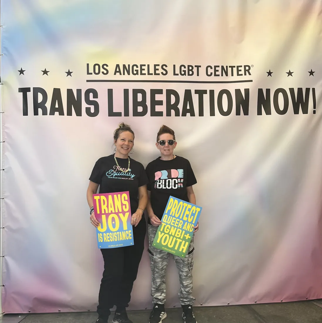 Jaki standing with a community member at an advocacy event, holding signs reading 'Trans Joy is Resistance' and 'Protect Queer and TGNBI+ Youth' beneath a 'Trans Liberation Now' banner.