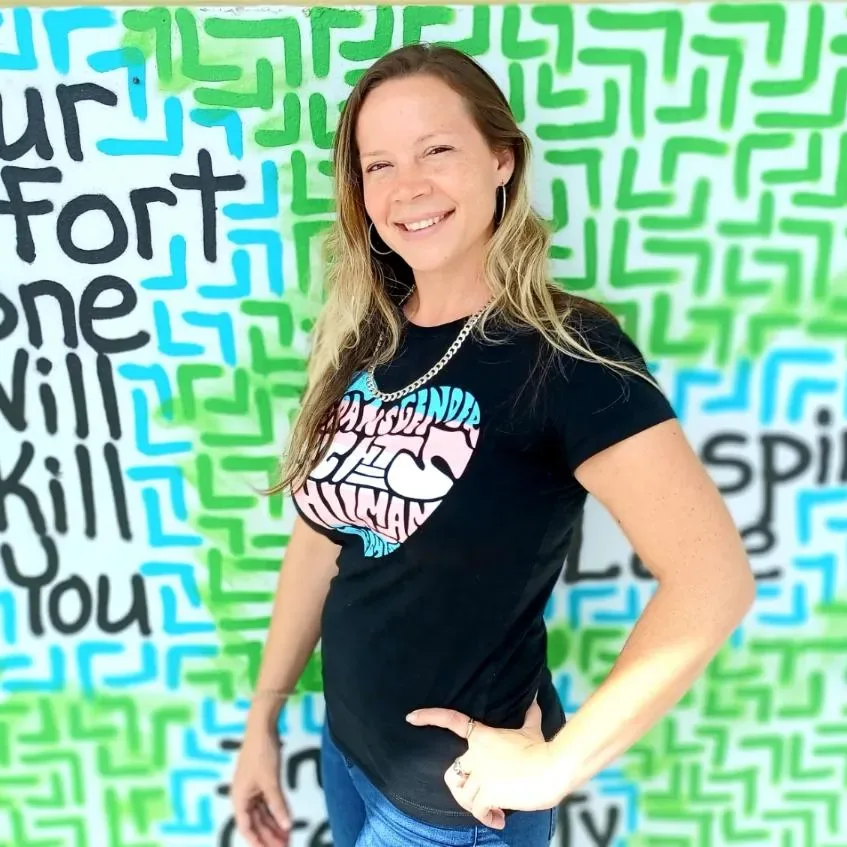 Jaki Neering smiling, standing in front of a vibrant green and blue mural, wearing a 'Transgender is Human' shirt.